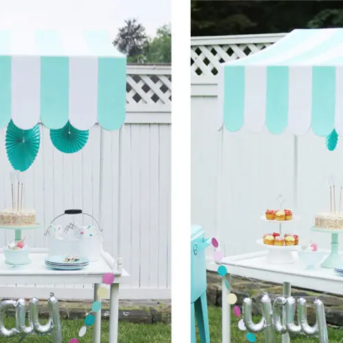 Outdoor dessert stand under a scalloped-edge blue and white striped canopy. Displays cakes, pastries on tiered stands, and cake under a glass dome, with turquoise decorations on the backdrop—perfect for those inspired by a DIY scallop table top canopy project.