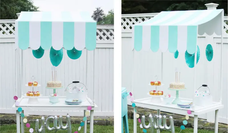 Outdoor dessert stand under a scalloped-edge blue and white striped canopy. Displays cakes, pastries on tiered stands, and cake under a glass dome, with turquoise decorations on the backdrop—perfect for those inspired by a DIY scallop table top canopy project.