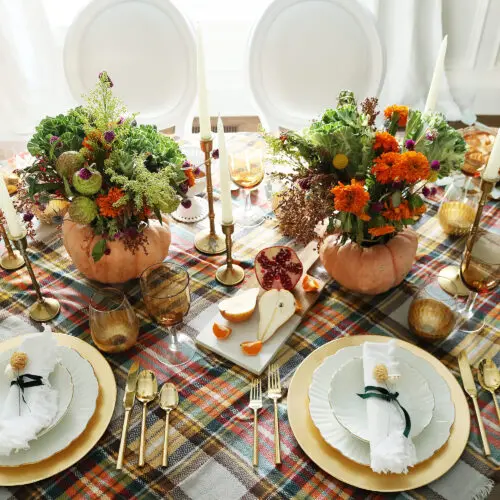 A festive table setting features plates, gold flatware, and goblets on a plaid tablecloth. Two pumpkin vases with colorful flowers serve as centerpieces, surrounded by candles and fruit.