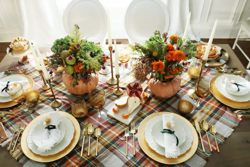 A festive table setting features plates, gold flatware, and goblets on a plaid tablecloth. Two pumpkin vases with colorful flowers serve as centerpieces, surrounded by candles and fruit.