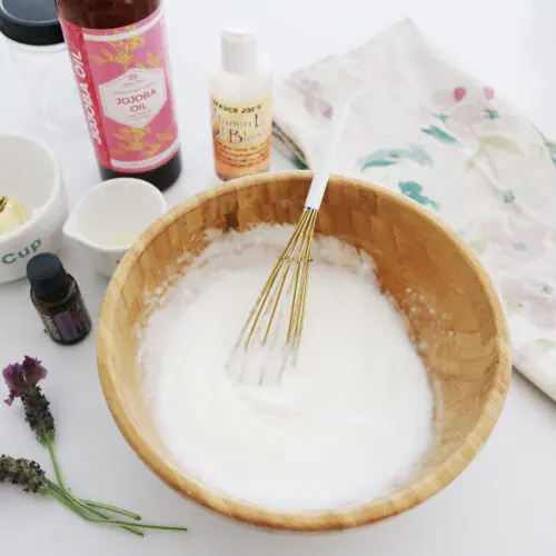 A wooden bowl containing a homemade cosmetic mixture with a whisk, surrounded by ingredients and floral decorations on a white surface.