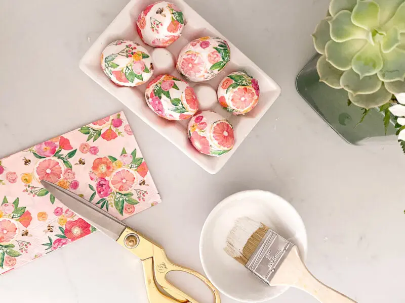 A crafting table featuring floral painted easter eggs in a carton, gold scissors, a paintbrush, and a napkin with a matching floral pattern, accompanied by a succulent plant.
