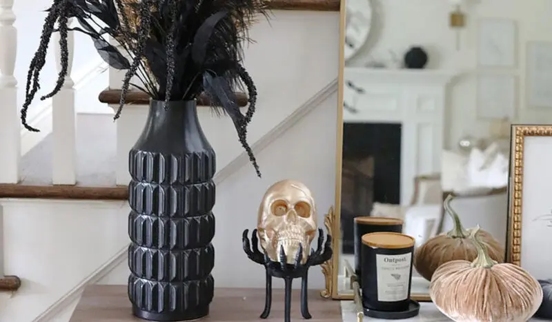 A halloween-themed mantel decor featuring a golden skull, black feathered branches in a vase, pumpkins, and a candle.