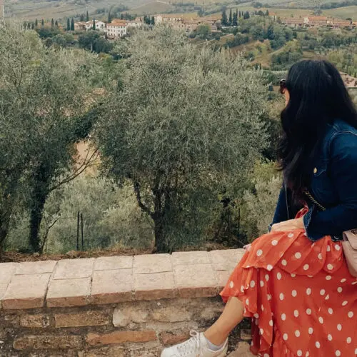 A woman in a red polka-dot dress and denim jacket sits on a stone wall, overlooking a scenic tuscan landscape with olive trees.