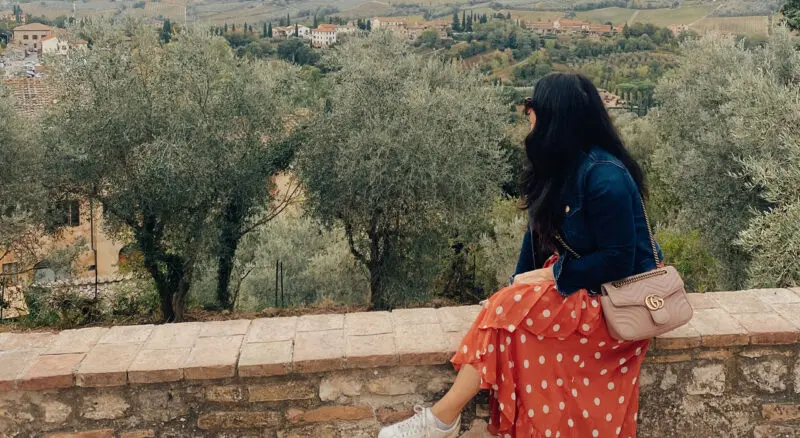 A woman in a red polka-dot dress and denim jacket sits on a stone wall, overlooking a scenic tuscan landscape with olive trees.