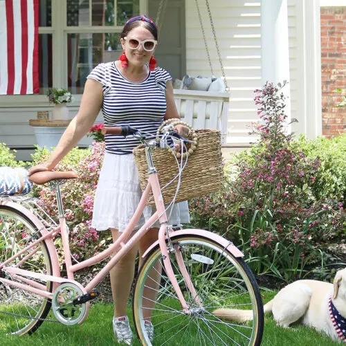A woman in a striped top and white skirt stands with a pink bicycle by a house, with a dog lying on the grass nearby.