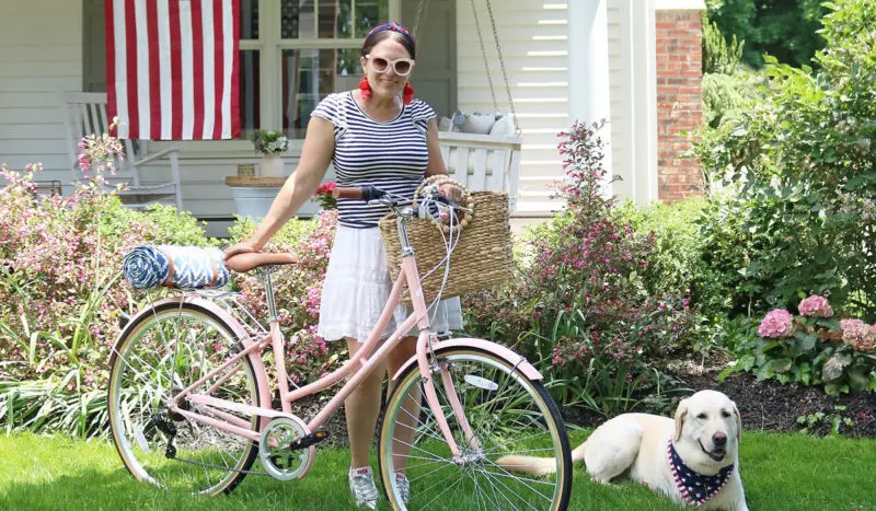 A woman in a striped top and white skirt stands with a pink bicycle by a house, with a dog lying on the grass nearby.
