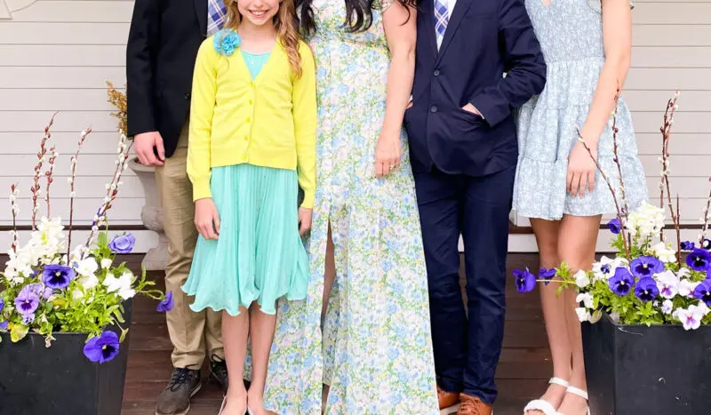 A family of five, with three children and two adults, posing together on a wooden porch, flanked by flower pots.