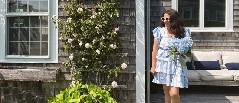 A woman in a blue and white dress smiling and holding a large bouquet of blue flowers next to a gray wooden house with climbing roses.