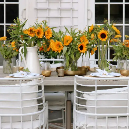 Outdoor dining setup with white chairs and wooden tables decorated with sunflowers in vases and white dinnerware.