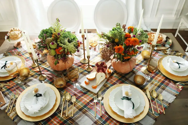 Elegant dining table set for a festive meal with floral arrangements in carved pumpkins, white plates, gold cutlery, and checked tablecloth.