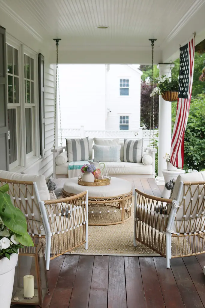 A cozy porch with a hanging swing bed, striped cushions, two wicker chairs, a round woven table, and an American flag hanging. The space includes plants and a candle on a wooden floor.