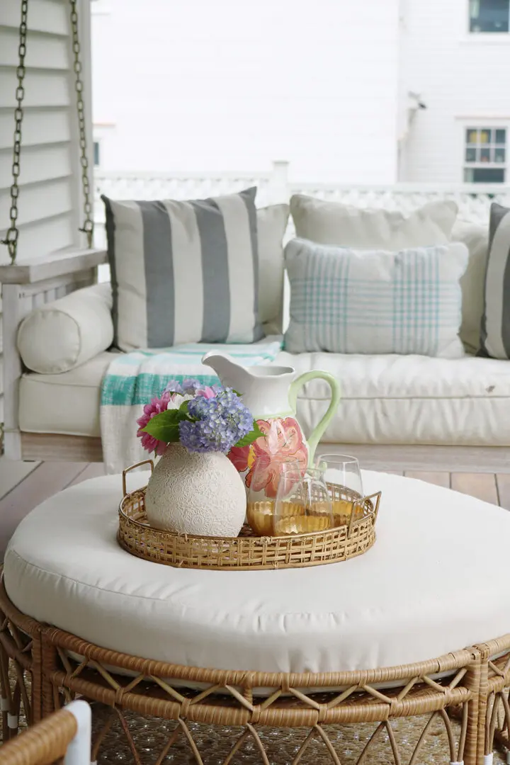 A tray with a vase of flowers, pitcher, and glasses is placed on a round ottoman in front of a cushioned outdoor sofa with striped and plaid pillows.