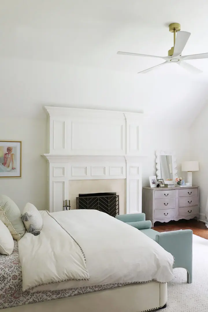 A bedroom with a white bed, light blue armchairs, and a grey dresser. A mirror and lamp sit on the dresser, and a framed picture is on the wall beside a white fireplace. A ceiling fan is overhead.