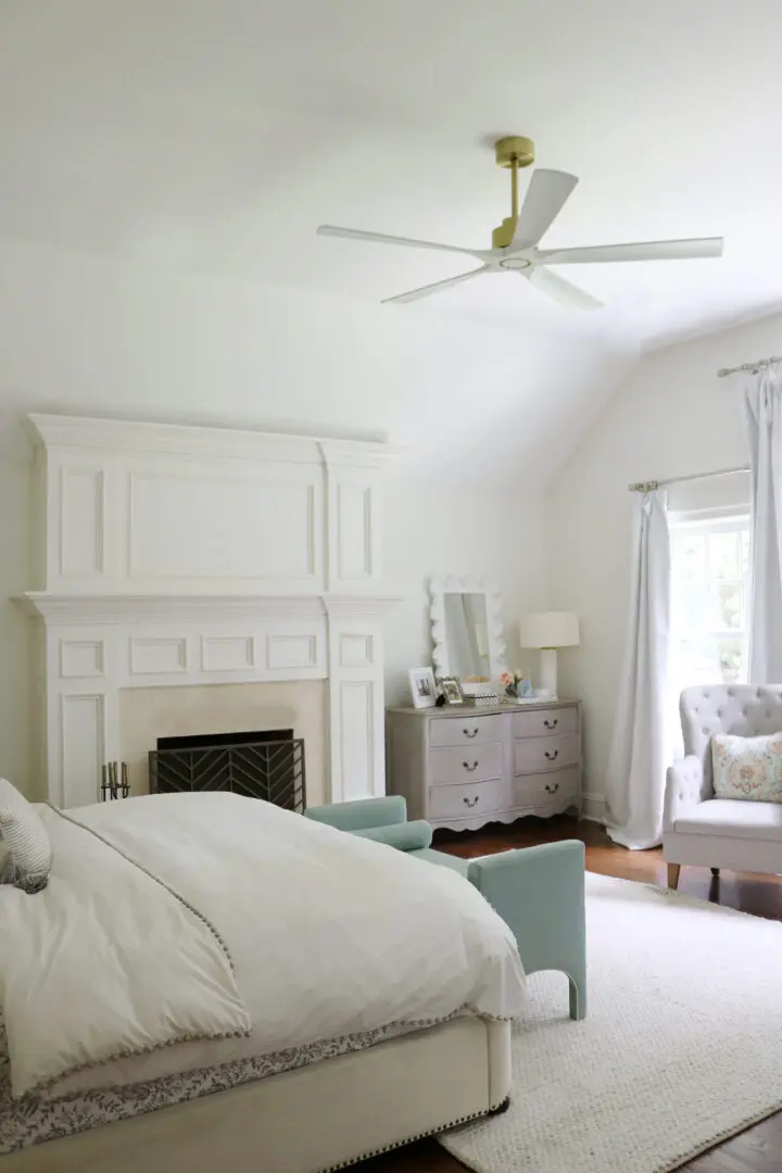 A bright bedroom with a white ceiling fan, a large white fireplace, and a gray upholstered armchair near a window. A bed with white bedding and a light blue bench are in the foreground. A dresser is next to the fireplace.