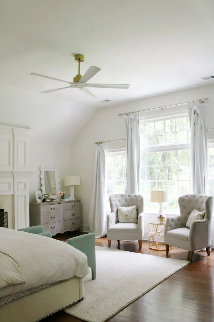 A bright bedroom with two armchairs by a window, a bed on the left, a dresser with a mirror, and lamps on nightstands. A ceiling fan is centered above. The room has a clean and airy feel.