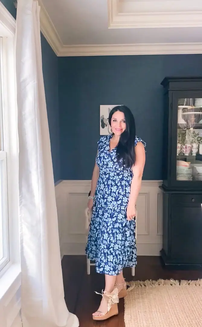 A woman stands smiling in a room with a dark wooden cabinet and a window with white curtains. She wears a blue floral dress and beige wedge sandals.