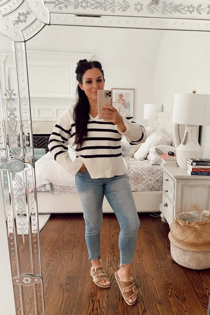 A person takes a mirror selfie wearing a white and black striped sweater, light blue jeans, and beige sandals in a bedroom with a neatly made bed and a lamp on a nightstand in the background.