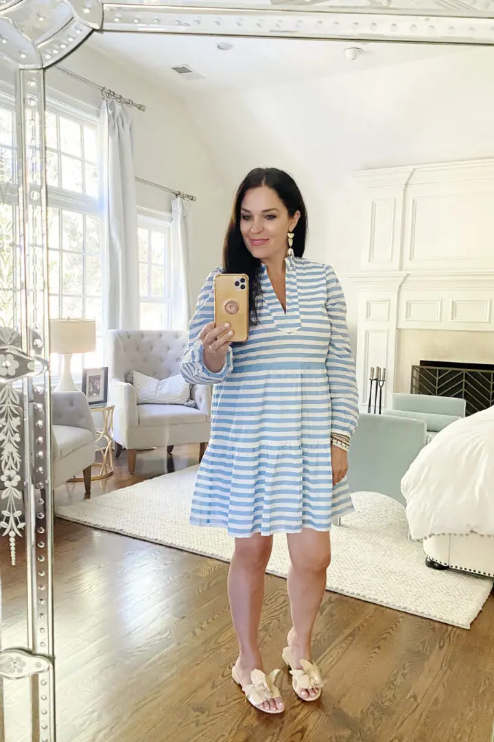 A woman in a blue and white striped dress takes a selfie in a mirror. She stands in a bright room with large windows, a white bed, and a fireplace in the background.