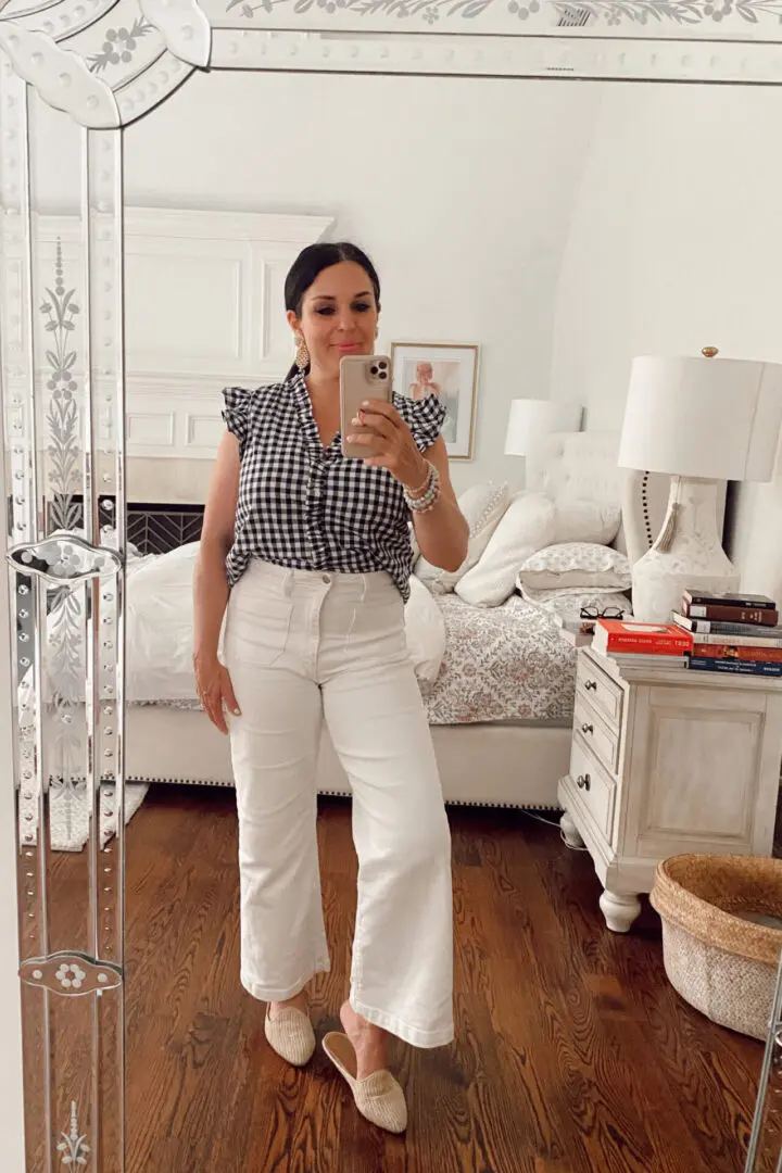 Woman stands in a bedroom taking a mirror selfie. She is wearing a navy and white checkered top and white pants. The room contains a bed, side table, and books.