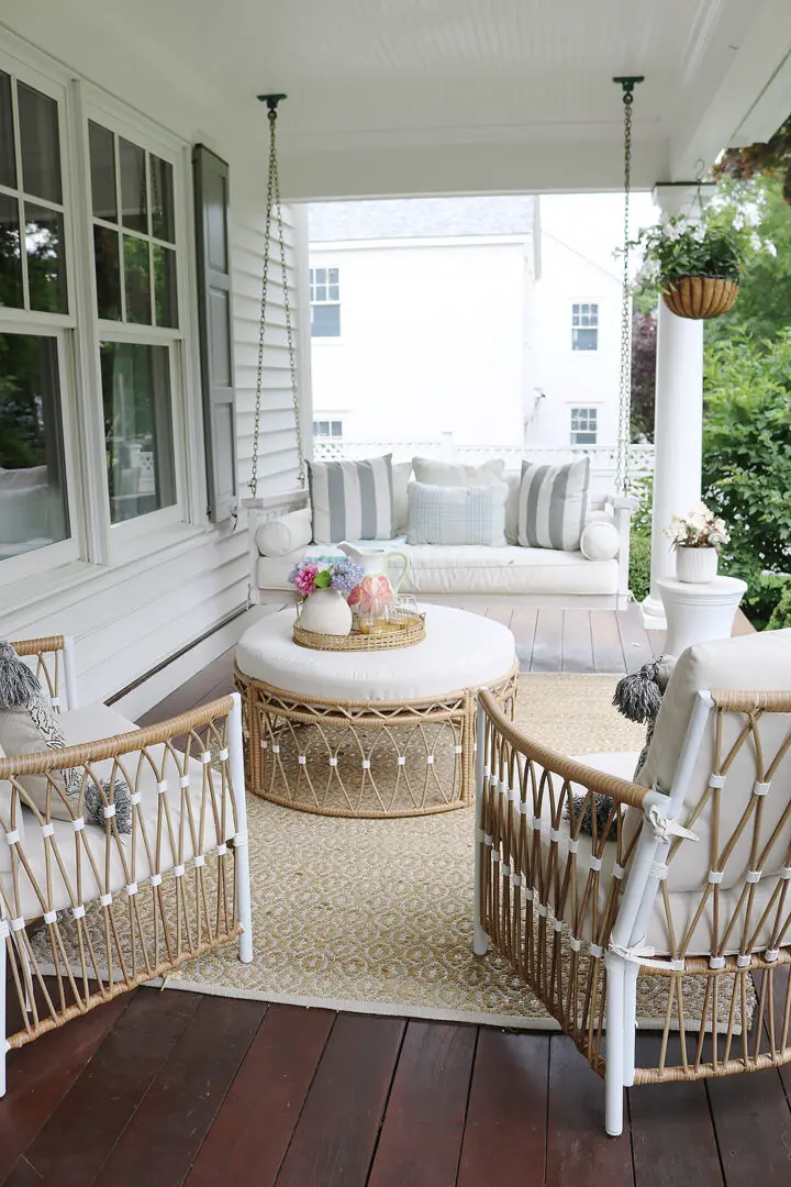 A covered porch features wicker furniture with white cushions, a round ottoman, and a wicker tray with flowers on a patterned rug. The porch overlooks a green yard and a white house.