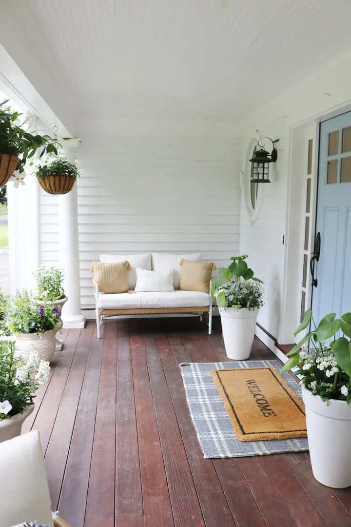A white porch with a light blue door features a white bench with tan pillows, potted plants, hanging baskets, and a "WELCOME" doormat on a plaid rug.