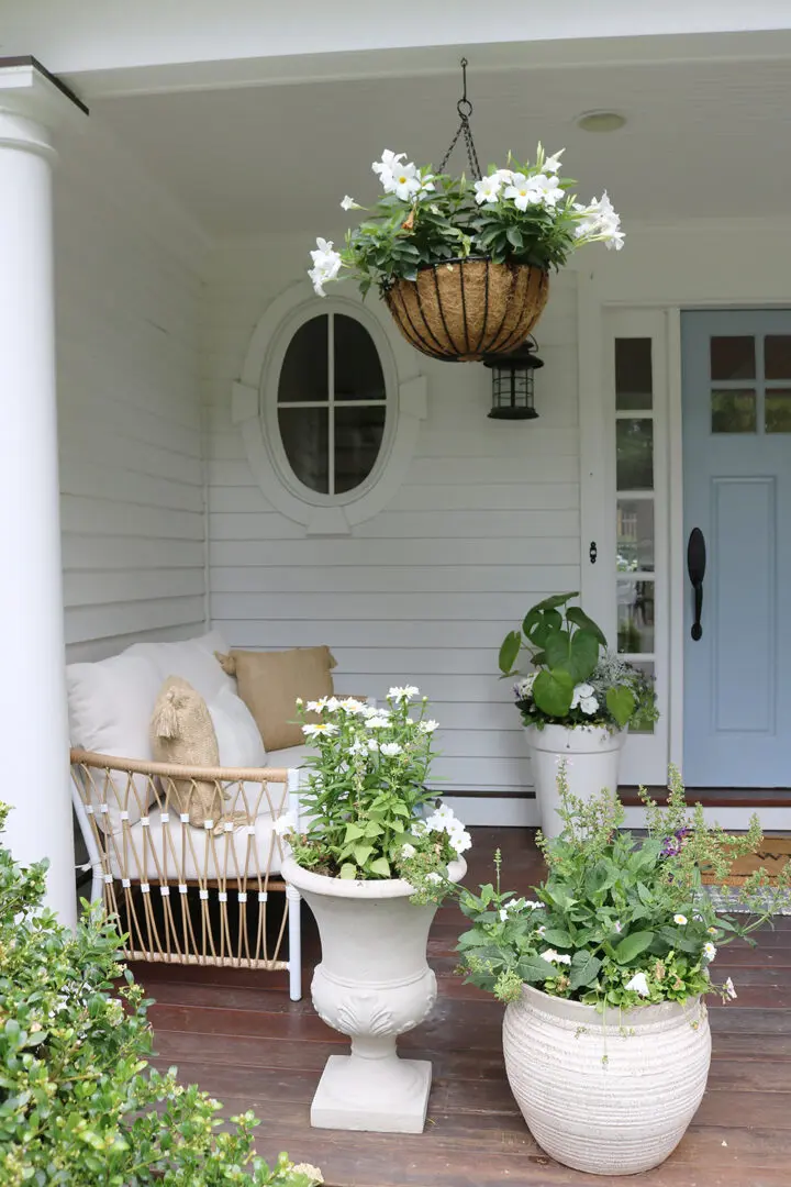 A front porch with white siding features a light blue door, a wicker chair with beige cushions, a hanging basket of flowers, and various potted plants.