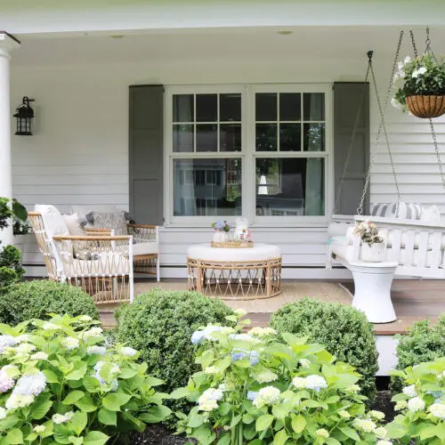 A porch with white columns features a charming white swing bench, a cozy wicker patio furniture set including chairs and a round table, hanging flower pots, and lush surrounding greenery.