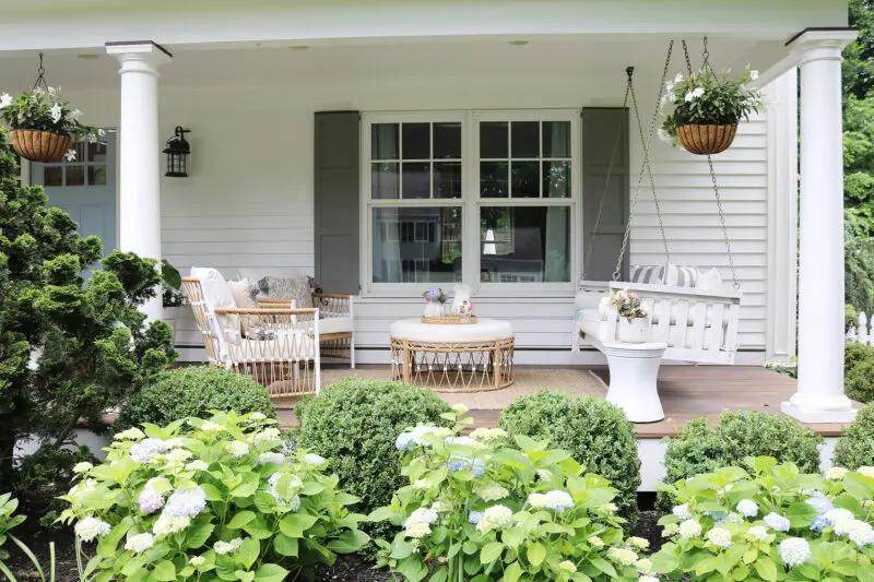 A porch with white columns features a charming white swing bench, a cozy wicker patio furniture set including chairs and a round table, hanging flower pots, and lush surrounding greenery.