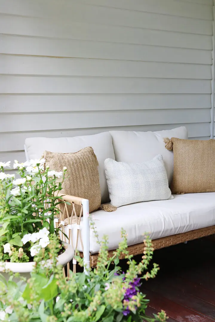 A white outdoor sofa with beige and white cushions sits on a wooden deck, surrounded by potted plants with green leaves and white flowers. The background features horizontal siding.