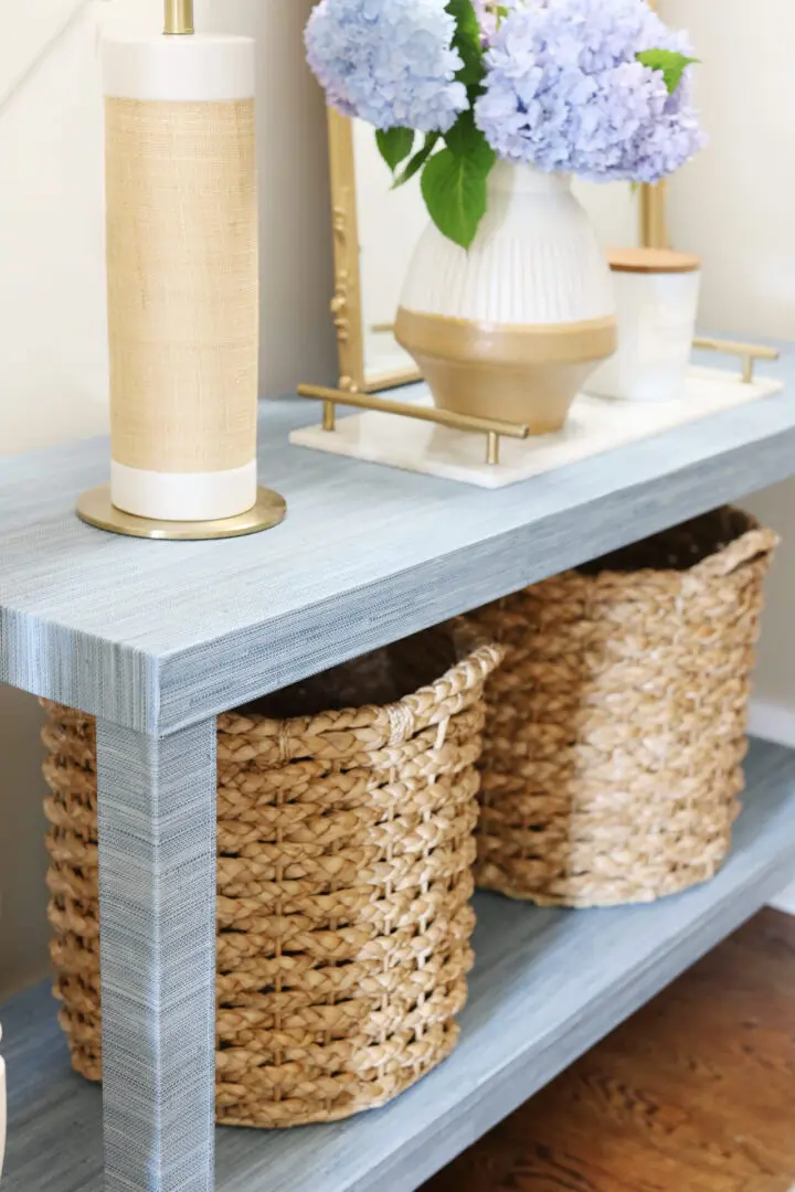 A blue wooden console table with two woven baskets on the lower shelf, a tall lamp, a vase with blue flowers, and other decor items on the top shelf.