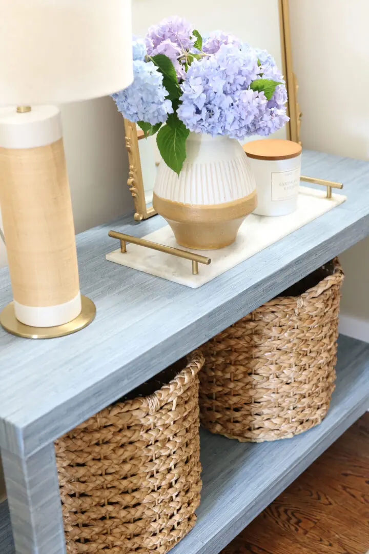 A light blue console table with a table lamp, a vase of blue and purple hydrangeas, a scented candle, and two woven baskets on the lower shelf.