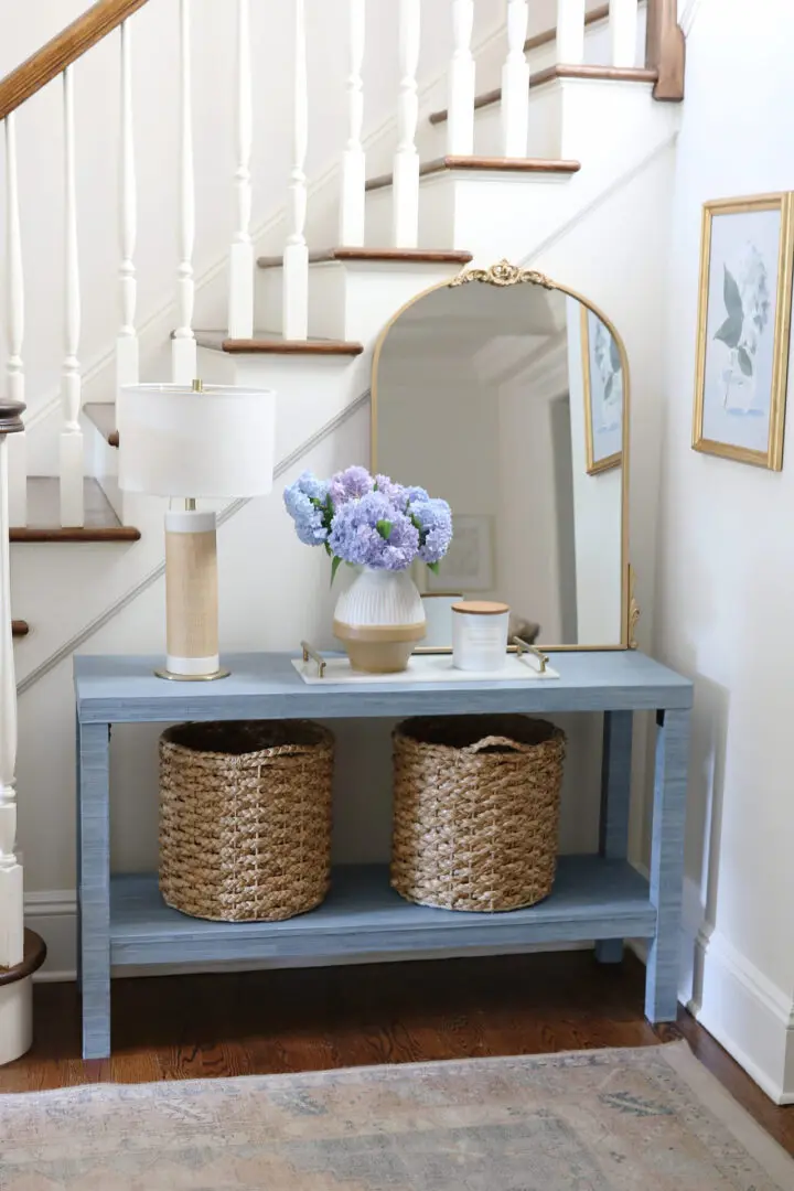 A staircase corner with a light blue console table, large mirror, table lamp, vase of purple flowers, and two woven baskets below. Two framed pictures are hung on the adjacent wall.