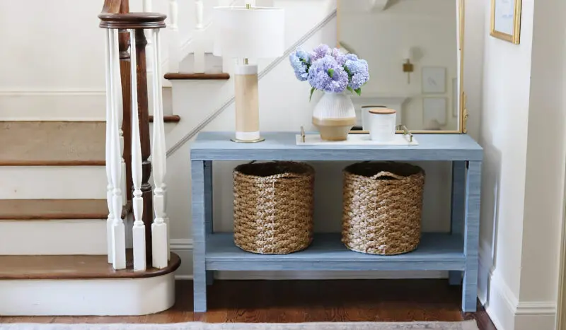 A hallway features a blue console table with two woven baskets underneath, a table lamp, a vase with hydrangeas, and a candle. The table is set against a staircase with white railings and wooden steps.