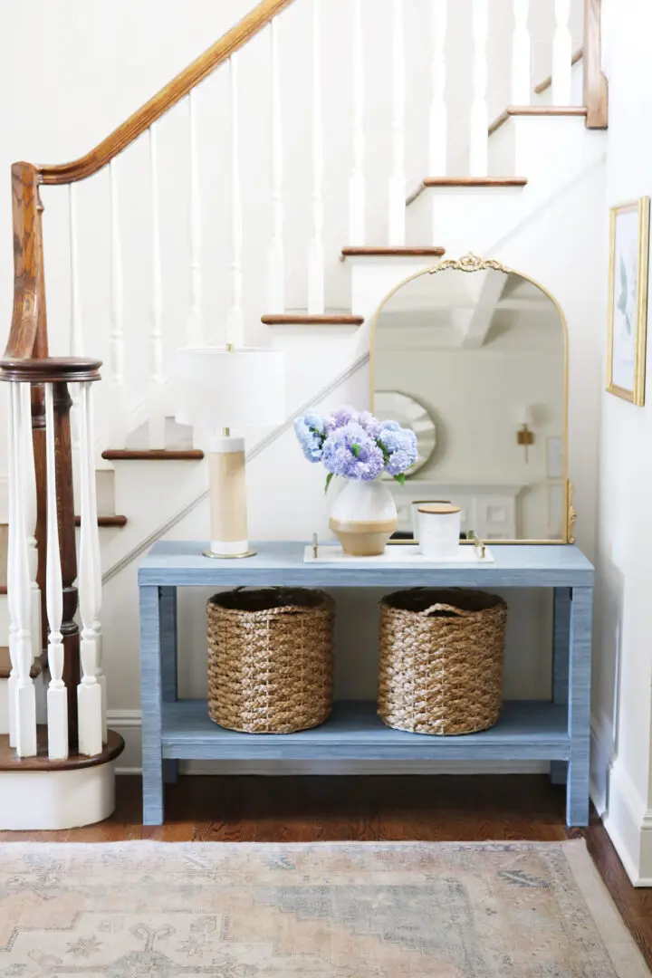 A blue console table with baskets, a lamp, and a vase of flowers is placed under a staircase with white balusters and a wooden handrail. An arched mirror leans against the wall above the table.