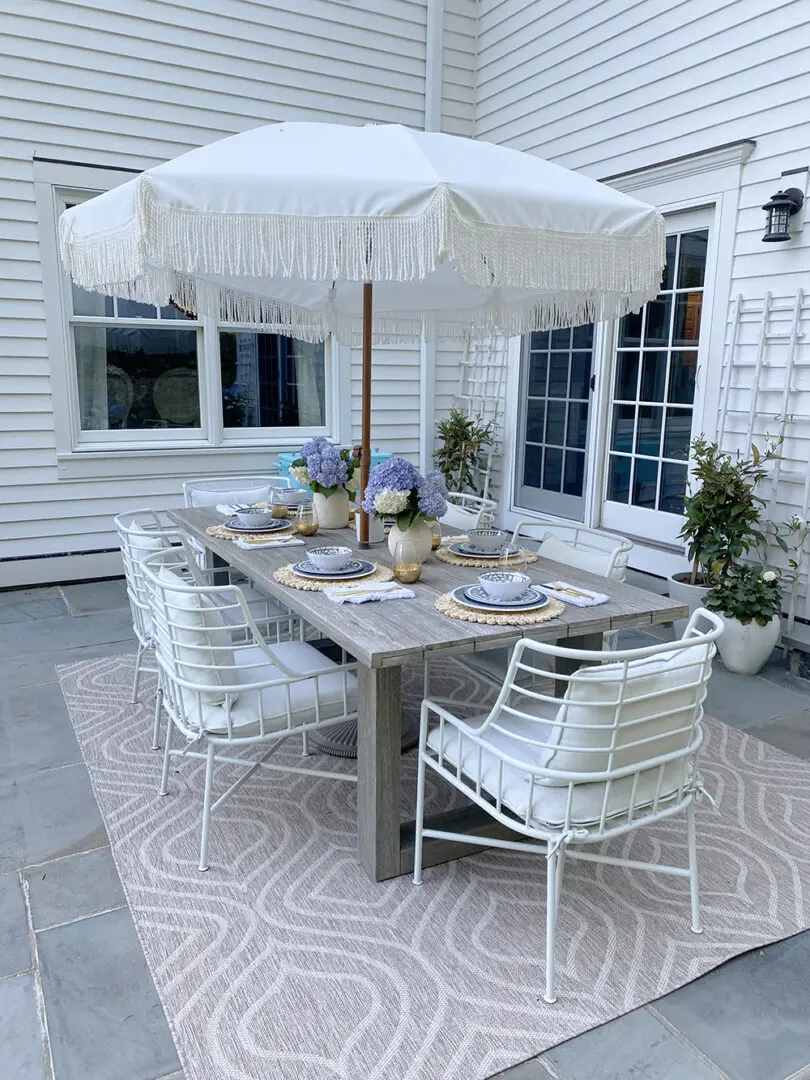 Outdoor patio dining set with a large fringed umbrella, a wooden table, white metal chairs with cushions, and hydrangea flower arrangements. The area is decorated with a patterned rug and potted plants.