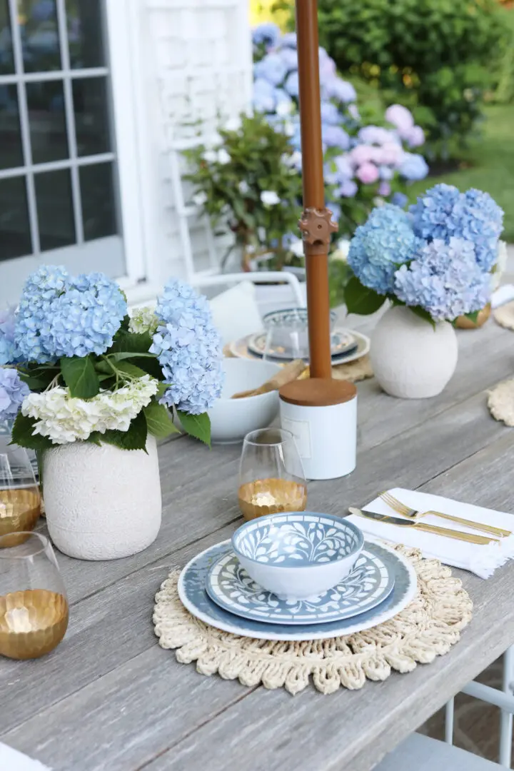 A wooden table is set for a meal with blue and white patterned dishes, cutlery, gold-rimmed glasses, and vases of blue hydrangeas.