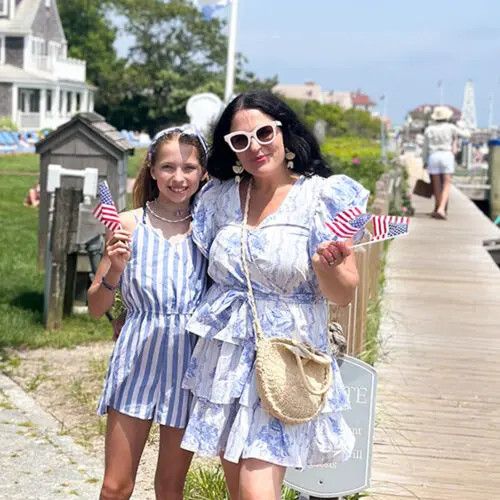 A woman and a girl stand on a boardwalk holding small American flags. They are dressed in light summer clothes and wear sunglasses. There are boats and houses in the background.