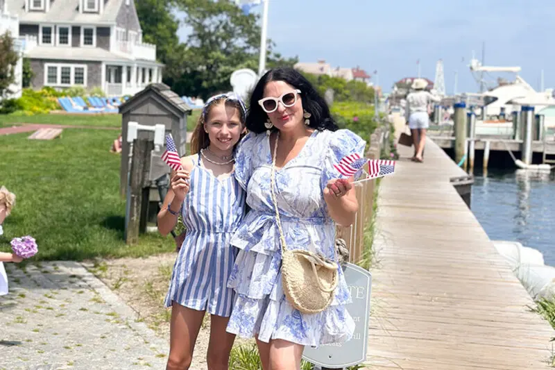 A woman and a girl stand on a boardwalk holding small American flags. They are dressed in light summer clothes and wear sunglasses. There are boats and houses in the background.