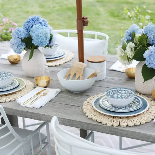 A wooden outdoor dining table is set with blue-and-white patterned dishes, gold-rimmed glasses, and vases of blue hydrangeas, surrounded by white chairs.