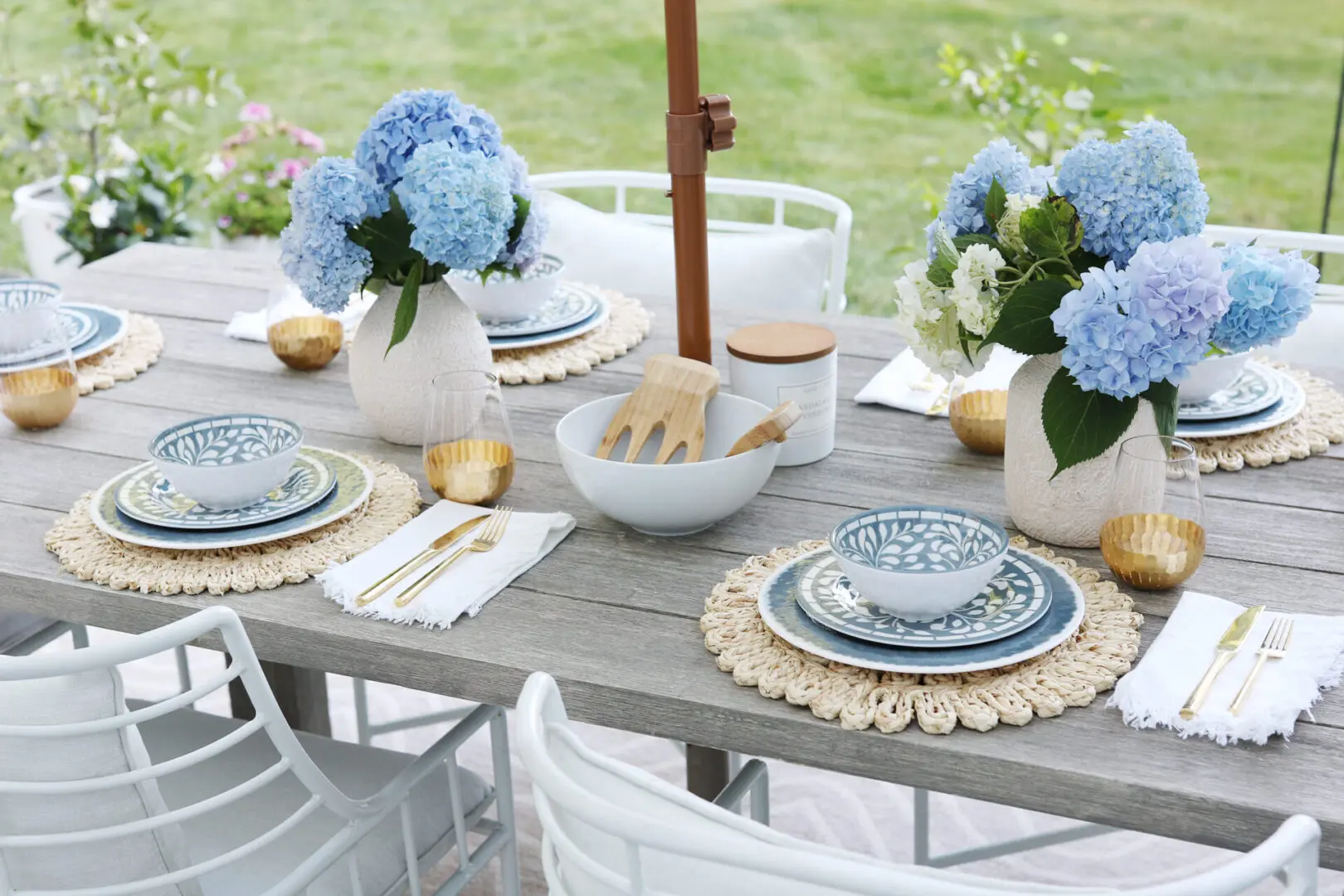 A wooden outdoor dining table is set with blue-and-white patterned dishes, gold-rimmed glasses, and vases of blue hydrangeas, surrounded by white chairs.