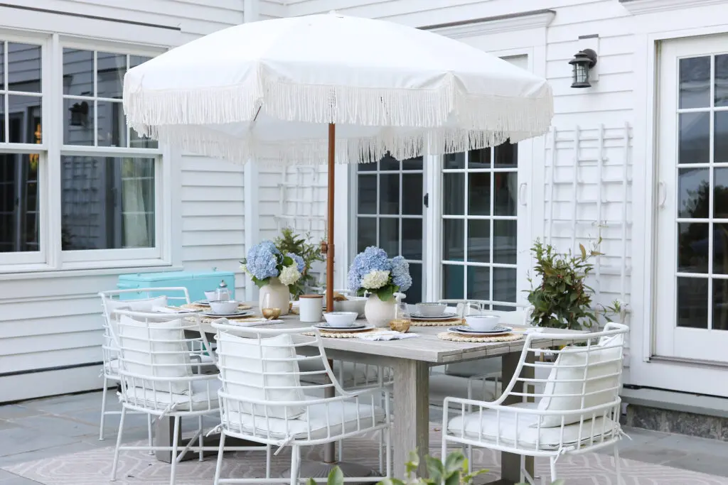 A patio dining area with a white table, six white chairs, and a large white umbrella. The table is set with dishware and decorated with blue hydrangea flowers.