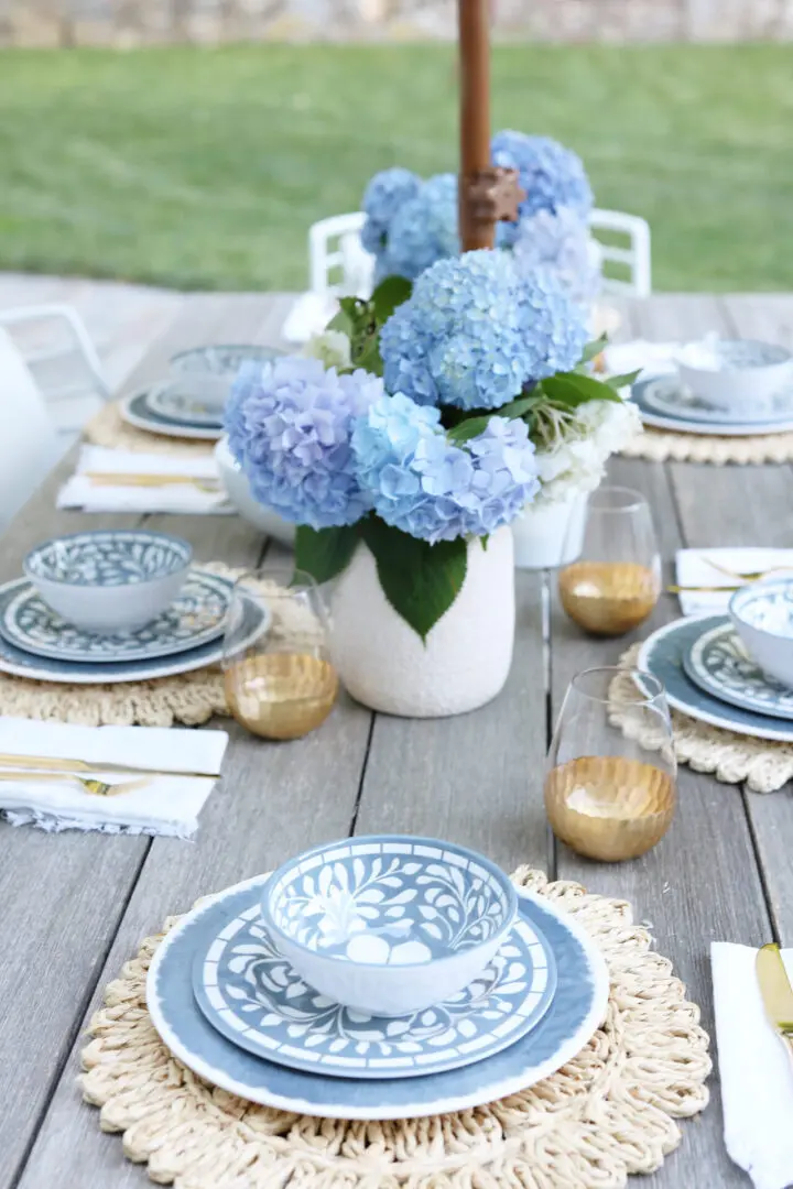 A wooden outdoor table set for six with blue and white patterned dinnerware, gold-rimmed glasses, white napkins with gold cutlery, and a vase of blue hydrangeas at the center.