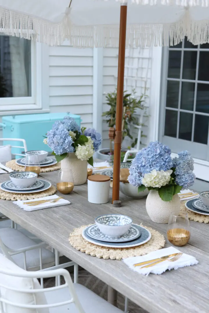 An outdoor dining table is set with blue and white plates, gold rimmed glasses, and vases of blue and white hydrangeas under a fringed umbrella. White chairs surround the table.