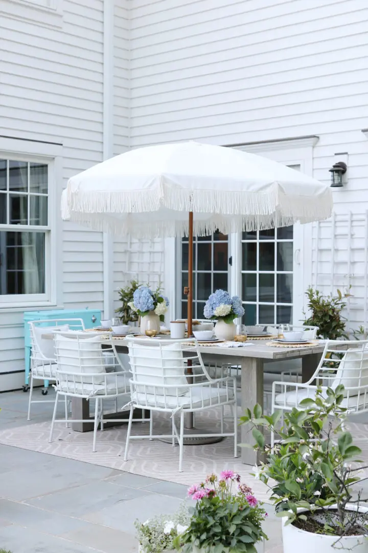 Outdoor patio with white table and chairs under a large white umbrella, set with plates and flowers. White siding, large window, and door in the background. Various green plants and flowers around.