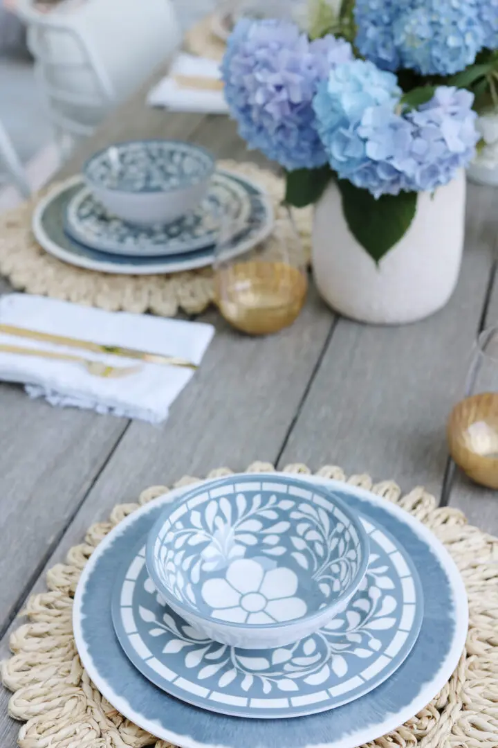 A neatly set dining table with blue floral patterned plates and bowls, gold utensils, and blue hydrangea centerpiece.