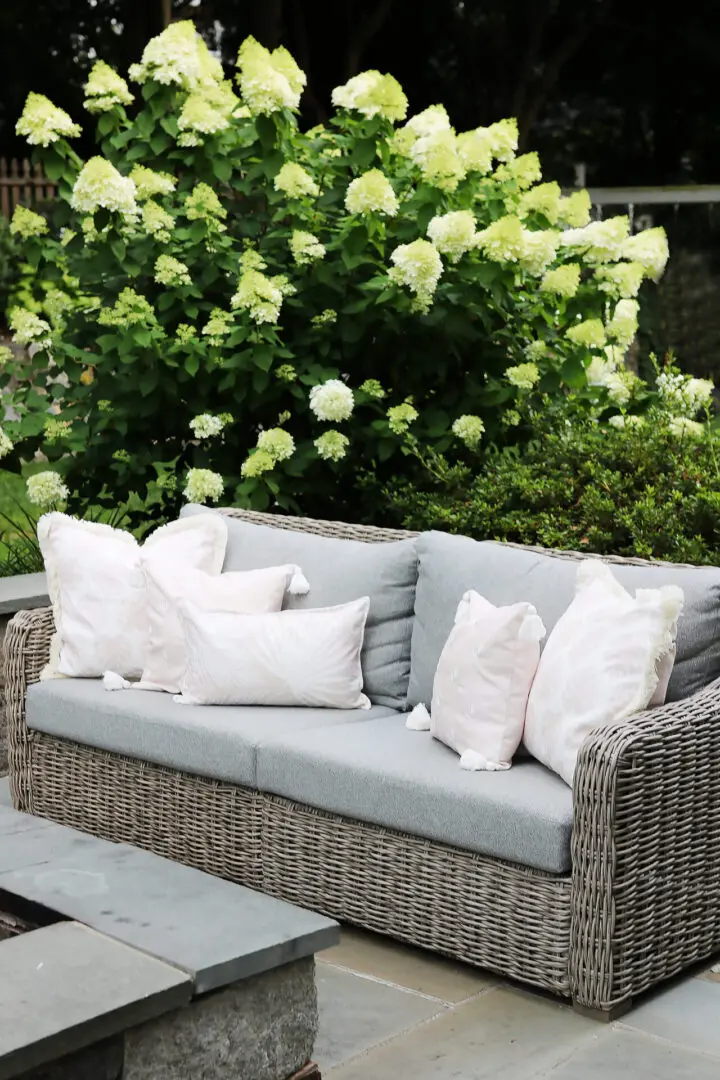 A wicker outdoor sofa with gray cushions and white patterned pillows is set in a garden. There are blooming hydrangea bushes and a stone table in the background.