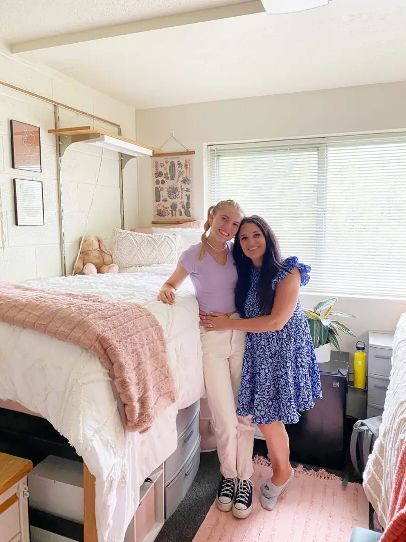 College Must-Have Essentials: The Ultimate Guide for Incoming Students Two people pose together in a well-lit room. One sits on a raised bed while the other stands beside. The room is sparsely decorated with a few personal items and a large window in the background.