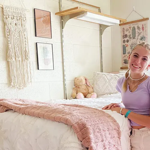 A young woman stands in a well-decorated bedroom with art on the walls, a teddy bear on the bed, and a window bringing in natural light. She smiles while leaning on the bed, which has a pink blanket.