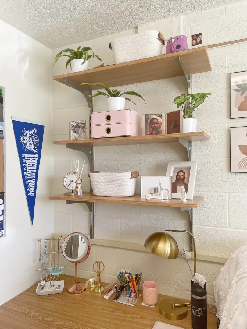 College Must-Have Essentials: The Ultimate Guide for Incoming Students A study desk with a lamp, mirror, and stationery is set against a wall. Above it are three wooden shelves holding decorative items, plants, and storage baskets. A blue pennant is pinned to the wall.